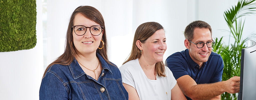 Frau steht vor einem Schreibtisch, vor ihr steht ein Bildschirm, ein Laptop und eine Tastatur. Sie schaut nach vorne und lächelt. An ihrem Schreibtisch stehen zwei weitere Personen und schauen auf den Bildschirm.