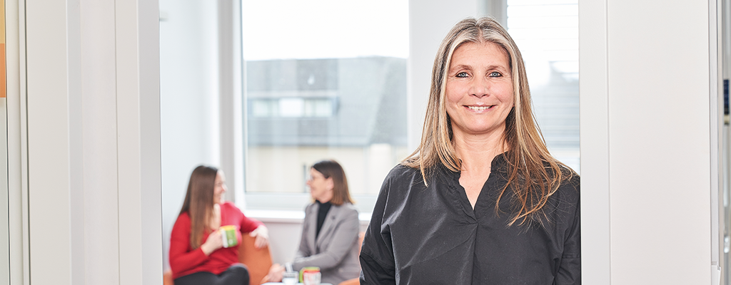 Frau steht in einem Türrahmen. Sie schaut nach vorne und lächelt. Hinter ihr im Raum sitzen zwei Personen auf Sesseln und unterhalten sich.