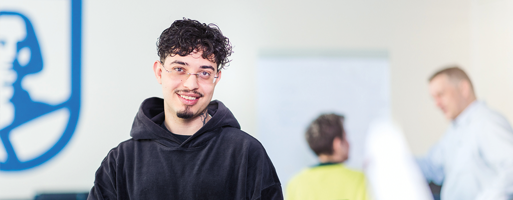 Junger Mann steht in einem Besprechungsraum, schaut nach vorne und lächelt. Im Hintergrund beschriften eine Frau und ein Mann ein Flipchart.