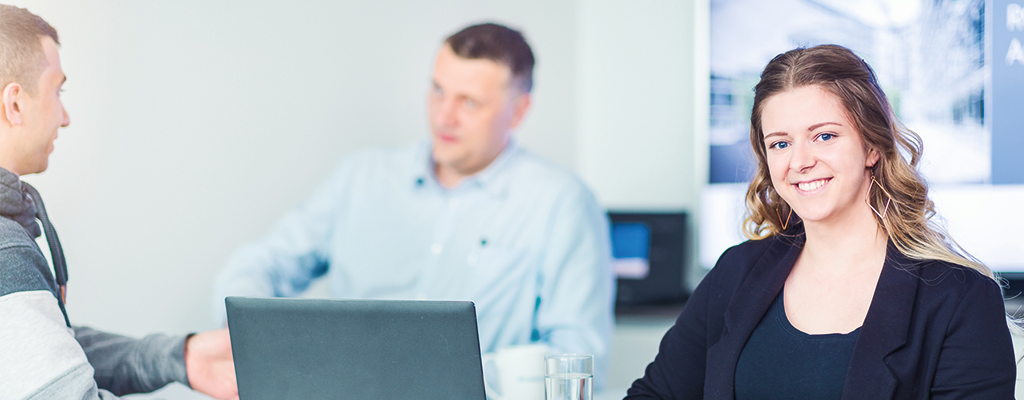 Junge Frau am Schreibtisch schaut nach vorne und lächelt. Im Hintergrund unterhalten sich zwei Männer.